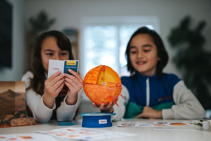 Two children sitting at a table, one holding a booklet on facts about the Sun, and the other holding an assembled papercraft template from Creative Park.