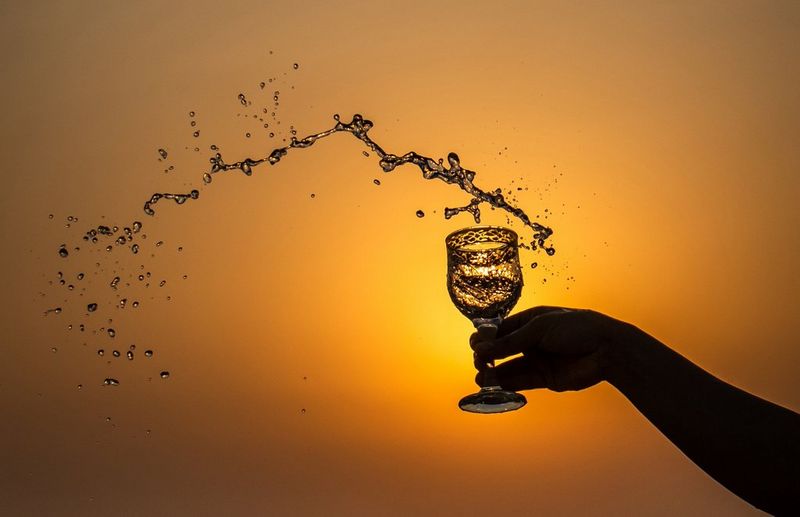 Droplets of liquid spray in an arc from a glass goblet held in a raised hand, silhouetted against an orange sky.
