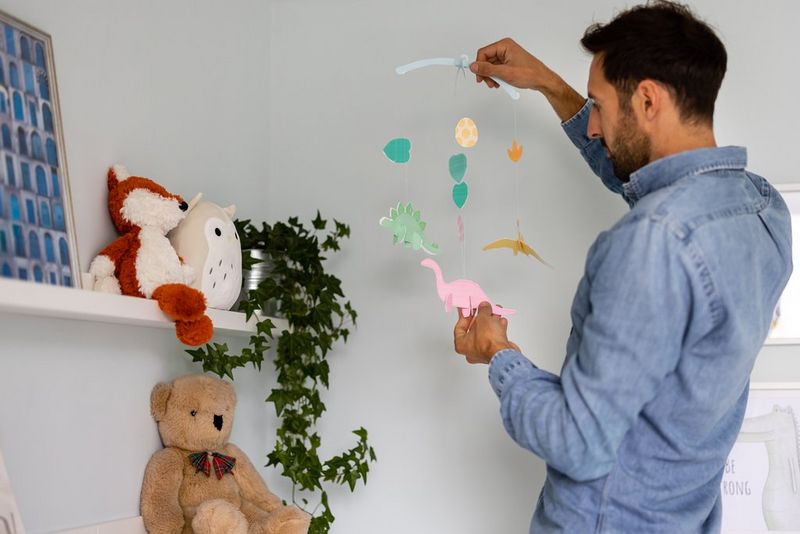 A man hanging a handmade 3D dinosaur mobile from the ceiling of a nursery.