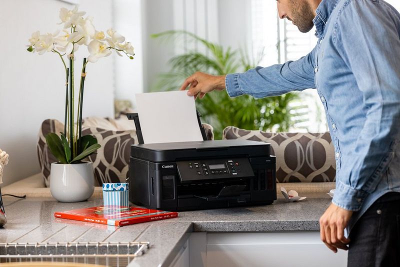 A man loading paper into a Canon PIXMA G6050 printer on a kitchen worktop.