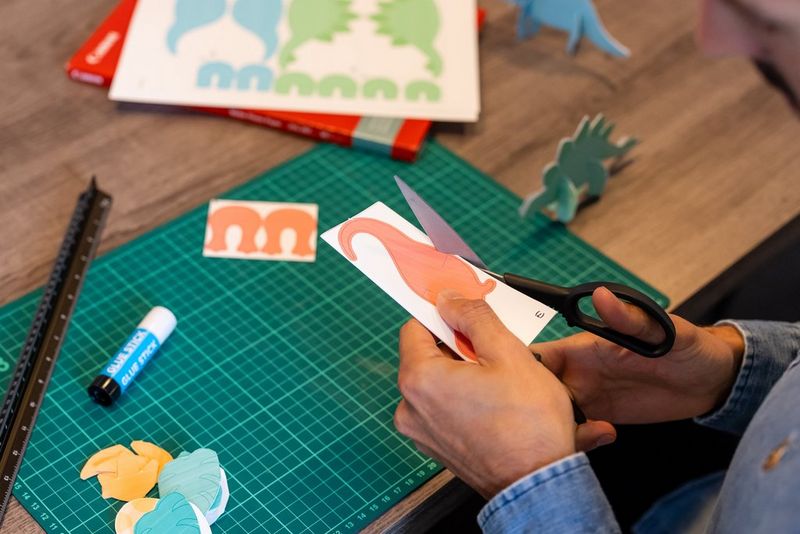 A man cuts out a picture of an orange dinosaur for a hanging mobile.