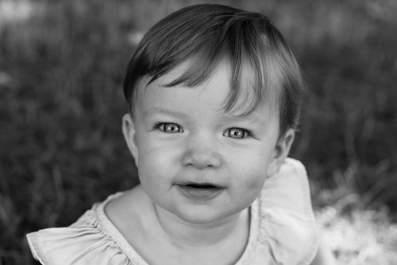 A black and white close-up portrait of a young child. She is staring intently at the camera and smiling.