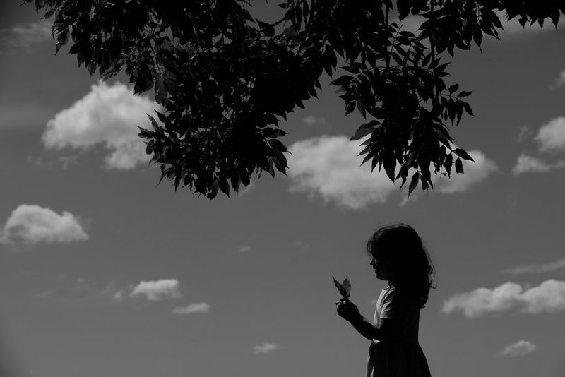 A black and white image of a young girl, cast in shadow, holding two leaves against a sky strewn with fluffy clouds.
