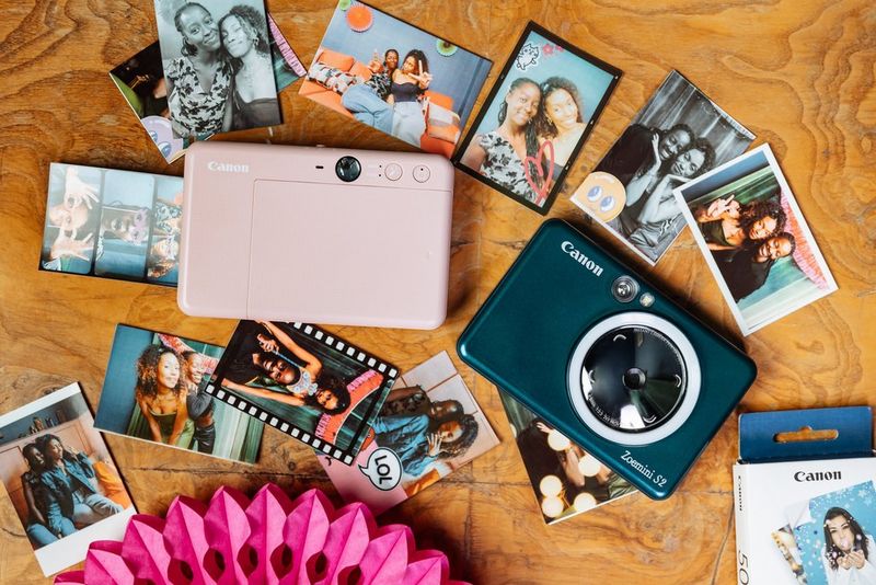 A flatlay showing two Canon Zoemini S2s – one rose gold and one teal – surrounded by several photo booth-style prints.