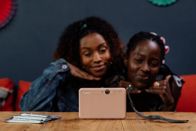 Two women smile and pose in front of a rose gold Canon Zoemini S2 sitting on a wooden table.