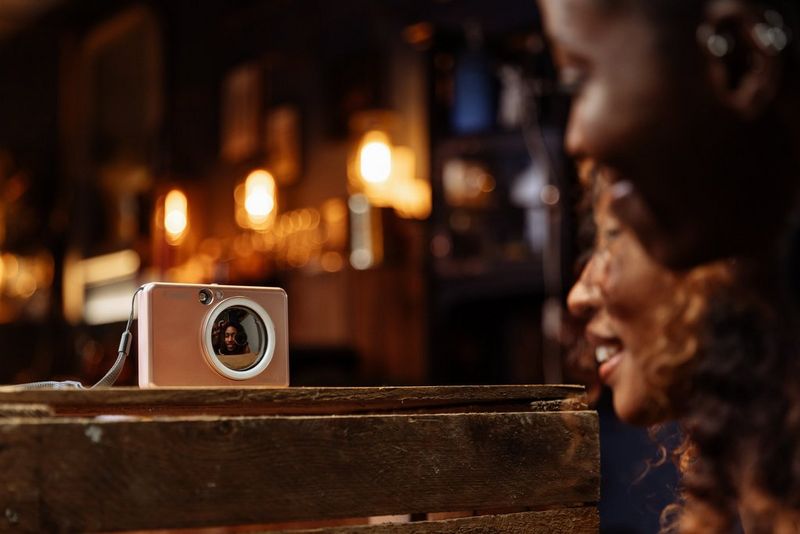 Two women smile and pose in front of a rose gold Canon Zoemini S2 sitting on a wooden table, their reflections visible in the Canon Zoemini S2's selfie mirror. 