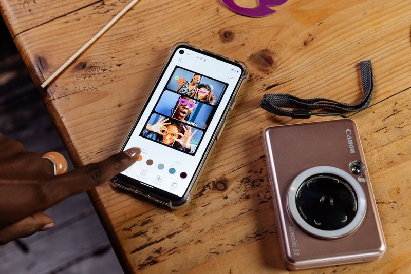 A person's finger edits an image collage on the Canon Mini Print app on a smartphone, next to a rose gold Canon Zoemini S2 lying on a wooden table. 