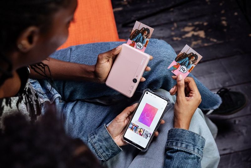 Two women sitting together, one holding a rose gold Canon Zoemini S2 in her hand, a print emerging from it, the other holding a smartphone showing the Canon Mini Print app. 