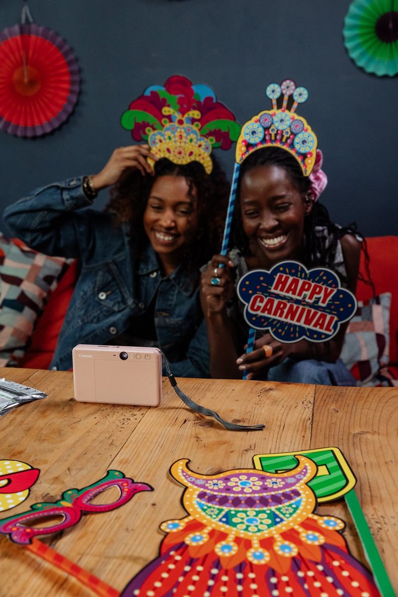 wo women pose with carnival-themed props in front of a rose gold Canon Zoemini S2 sitting on a wooden table. Other carnival-themed props lay on the wooden table. 