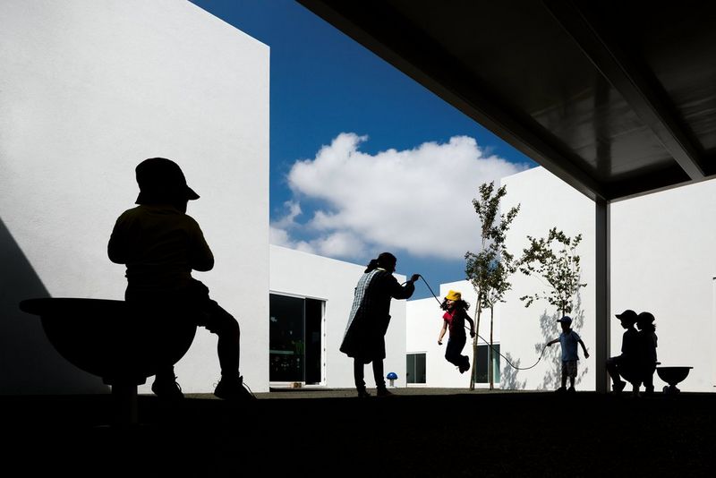 A woman and several children play with a skipping rope in the sunshine next to a stark white building. All but one of the children are seen in silhouette.