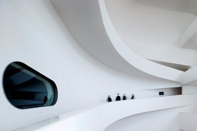 Four people are blurred in motion as they walk along a large corridor inside a huge white building with futuristic windows and walls.