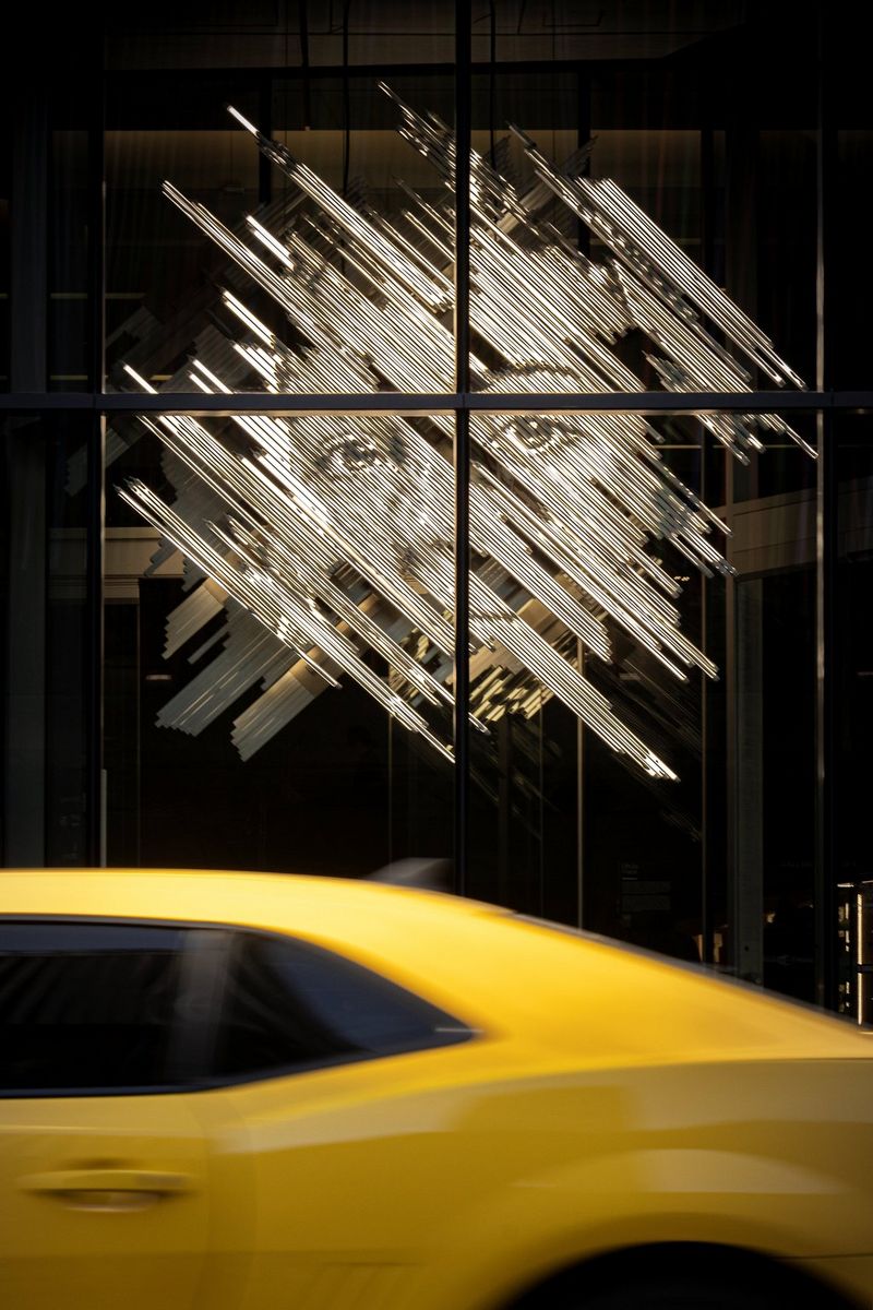 A large light installation featuring the face of a young woman can be seen through the window of a building as a yellow car drives by.