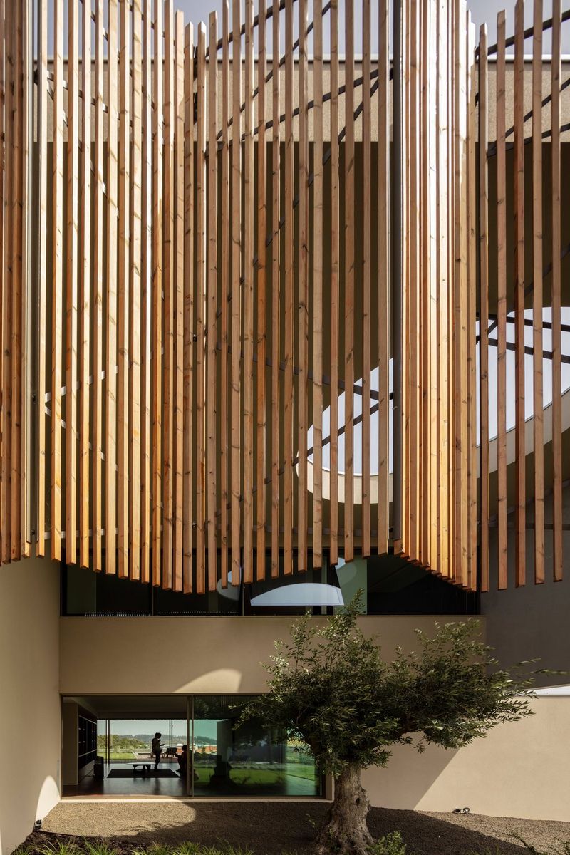Huge wooden slats hang vertically from a large building painted dark cream. In a room on the ground floor of the building, two people can be seen in silhouette.