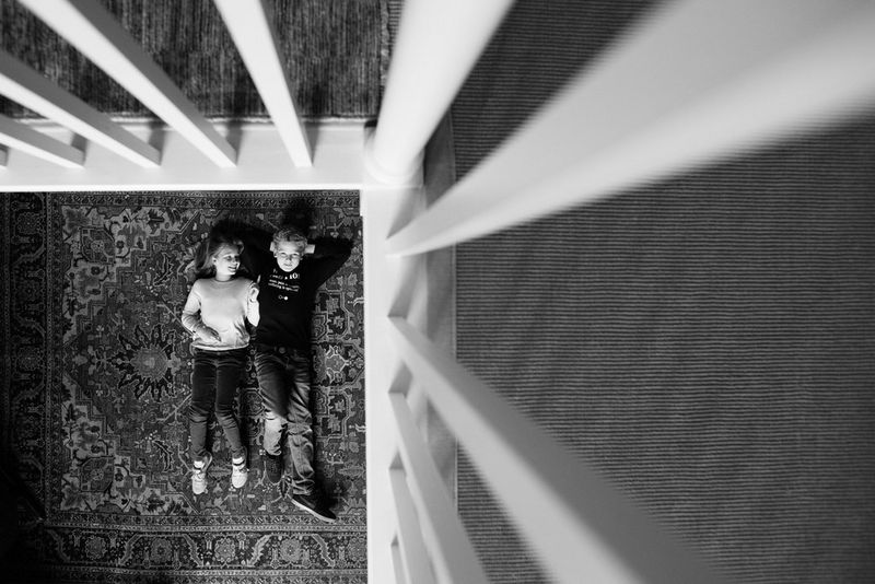 A top-down black and white shot, taken from the landing above, of two children lying on a rug framed by the banisters of the stairs.