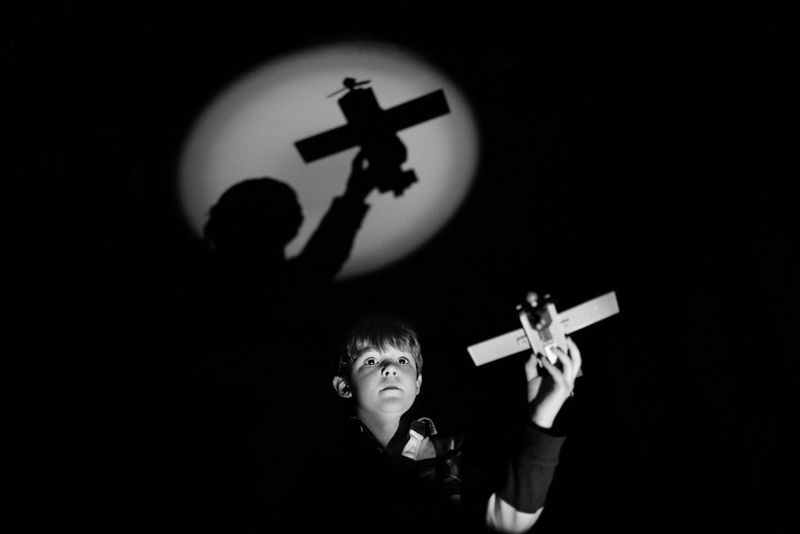 A little boy playing with a toy plane in a dark room. The shadow of the plane is reflected on the ceiling by a spotlight.