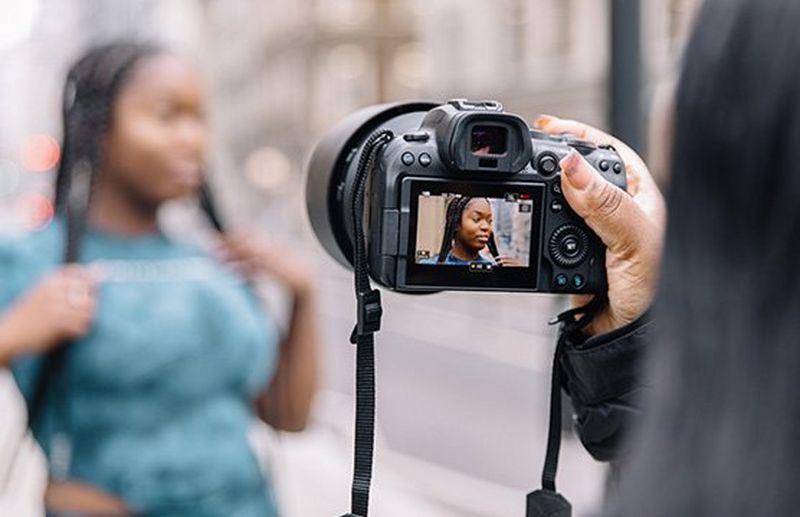A woman with long braids being photographed in the street by her friend, who is holding a Canon EOS R6.