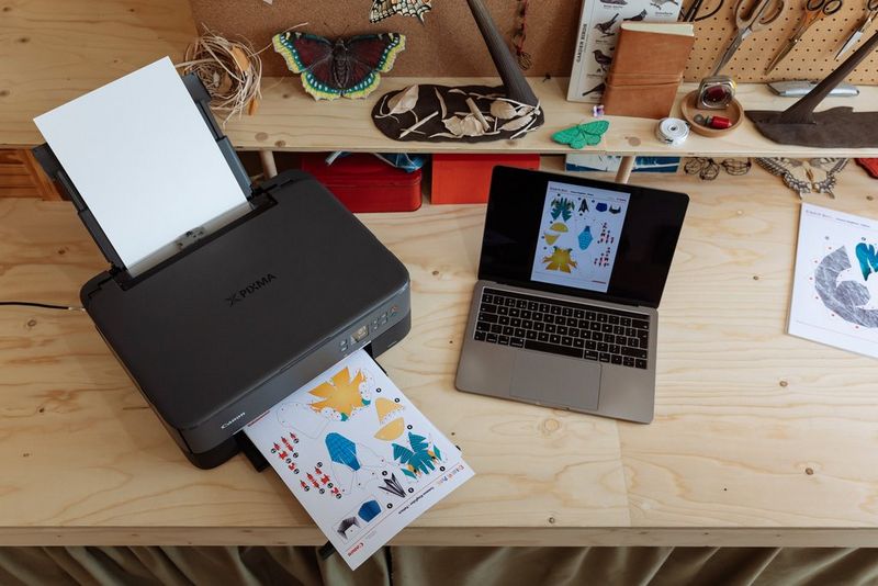 A Canon PIXMA printer sits on a worktop next to an open laptop, printing out a Canon Creative Park template that is shown on the laptop screen.