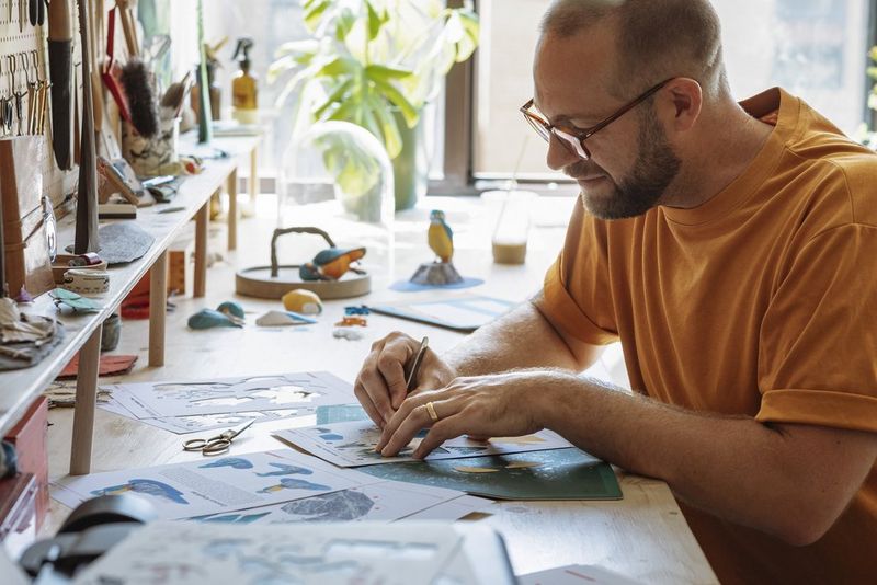 A man leans over a desk, carefully using a scalpel to cut out templates from sheets of paper on a cutting mat.