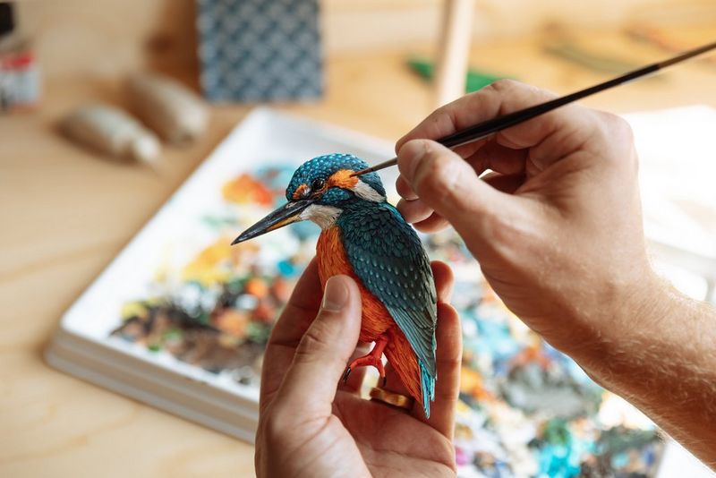 A person's hand holds a papercraft kingfisher while the other hand uses a thin brush to paint the kingfisher's head.