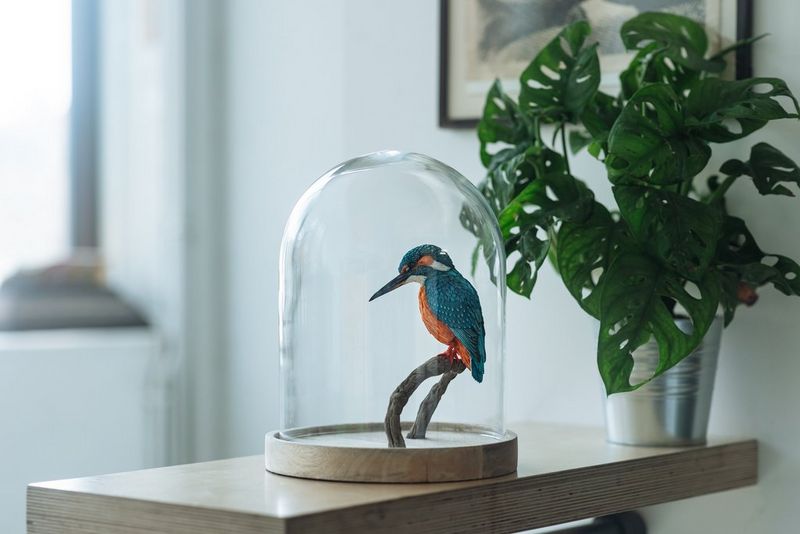 A finished papercraft kingfisher perches on a branch in a bell jar which sits next to a potted plant on a table.