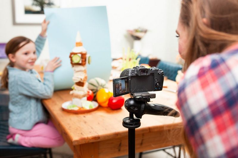 A little girl holding up a blue piece of paper behind a tower of food shaped like Big Ben. 
