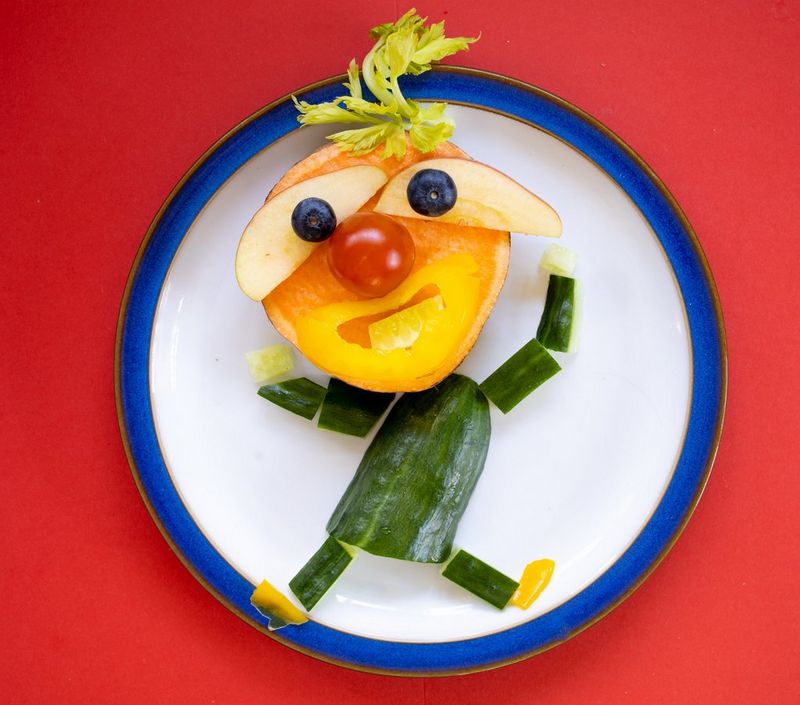 A person made of food, including cucumber, orange and apple, on a white plate. 