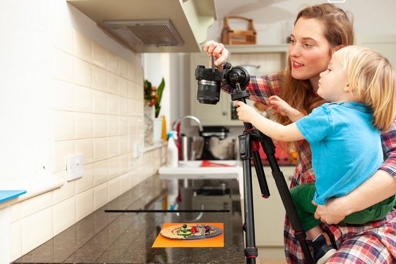 A mother holding her child as they set up a camera on a tripod to shoot a plate of food made into a face from above. 
