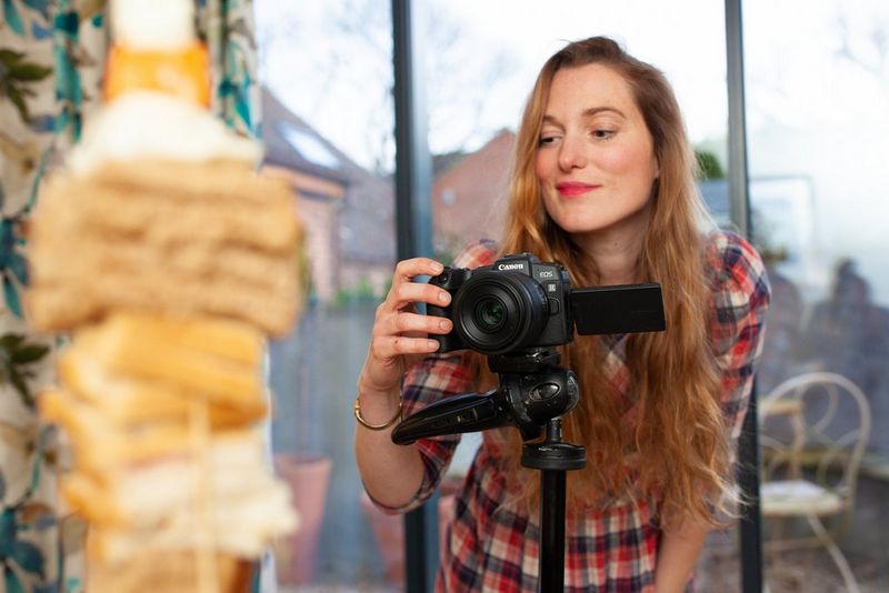 A woman photographing a food tower with a camera on a tripod by a large window. 
