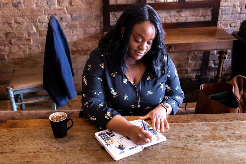 A woman sits at a wooden desk writing in a journal, a hot drink next to her.