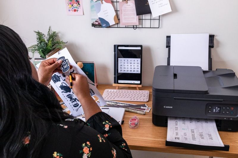 A woman uses a pair of scissors to cut out themed motifs downloaded from Creative Park. A Canon PIXMA printer sits on the desk. 