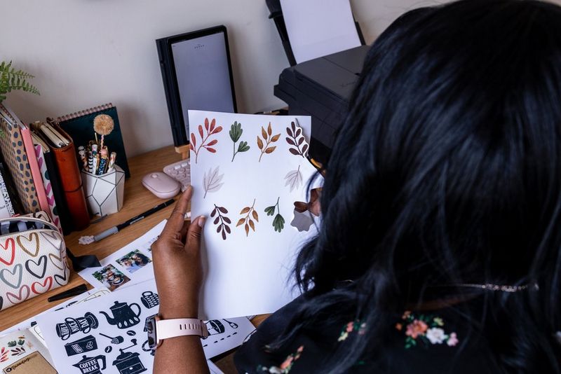 A woman cuts out a leaf illustration while sitting at a desk. On the desk is a Canon printer, notebooks and pens.