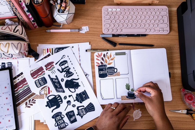 A pair of hands placing coffee-related stickers from Canon Creative Park into a journal. Next to them on the desk are various printed designs, a pink keyboard and items of stationery.