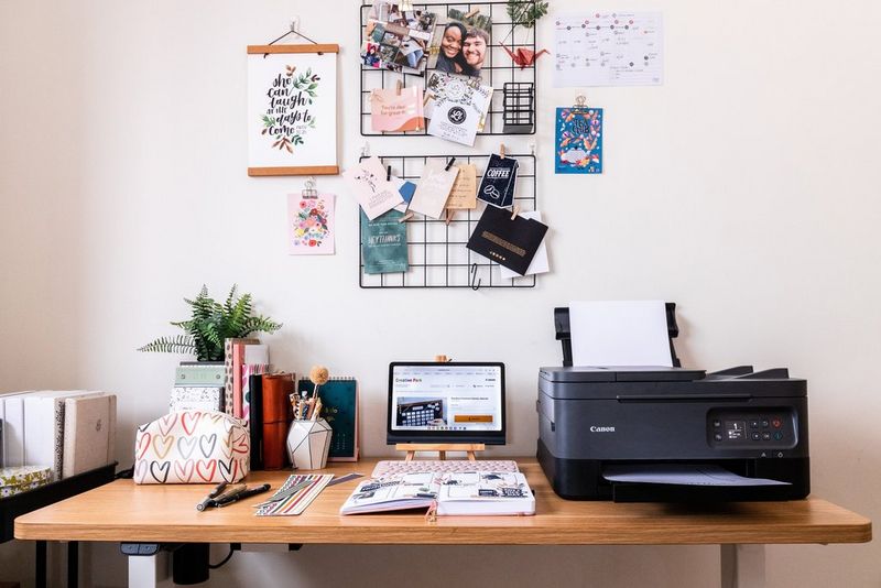 A Canon printer, tablet and various items of stationery on a wooden desk. Above the desk on the wall, postcards and notes are attached to a metal wall hanger. 