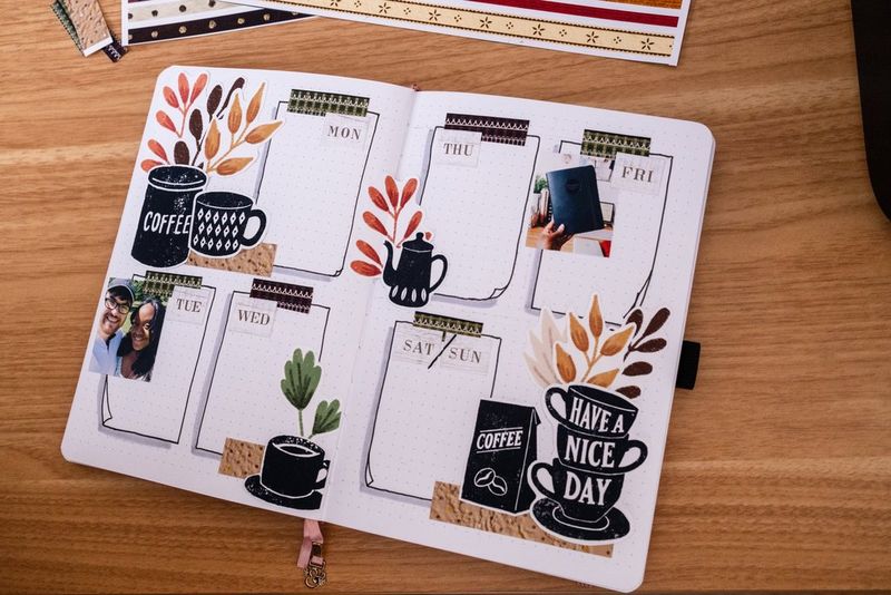 A top-down image of a wooden desk with a bullet journal open displaying daily to-do lists and leaf and mug motifs.