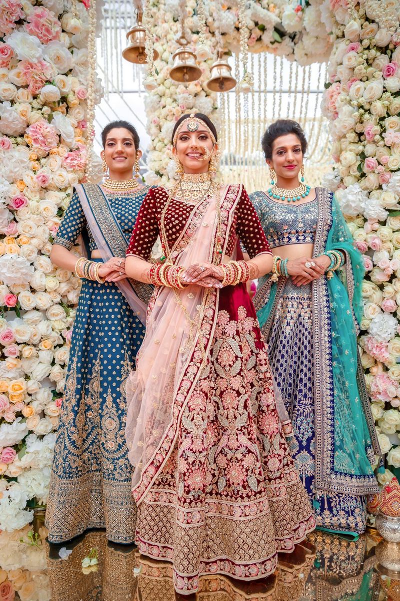 A group portrait of three women in brightly coloured traditional Indian dress, with henna and numerous bracelets on their arms, standing in front of a floral display.
