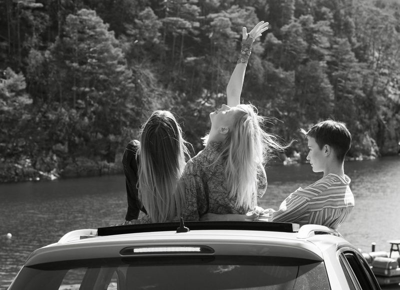 A black and white group portrait of three young women sitting on the bonnet of a car in front of a picturesque lake.