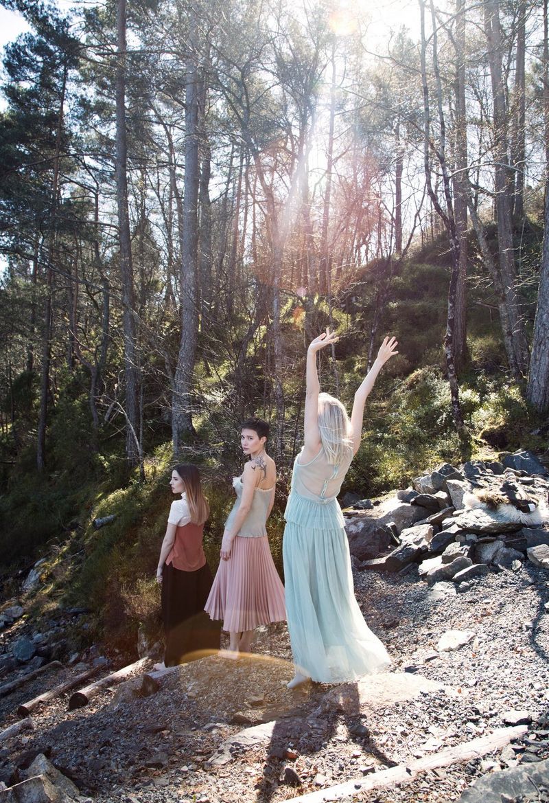Three young women in summer clothing walk through a forest as the sun shines through the trees. One of the women has her hands in the air.