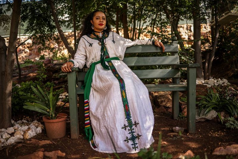 A woman wearing a long white dress and eye-catching jewellery sits on a bench outside her Johannesburg home.