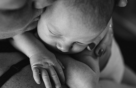 A black and white close-up shot of a mother kissing a sleeping baby she is holding to her shoulder.