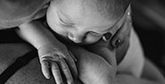 A black and white close-up shot of a mother kissing a sleeping baby she is holding to her shoulder.