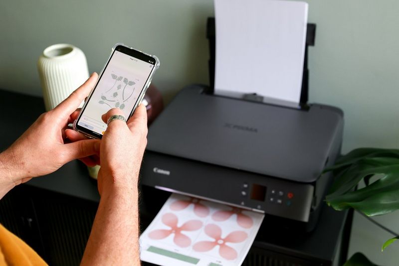 A pair of hands holds a smartphone with flower templates from Canon Creative Park on the screen, with petal templates emerging from a Canon PIXMA printer below. 