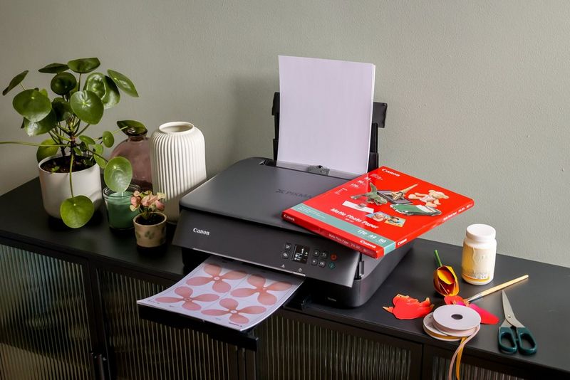 A print emerges from a Canon PIXMA printer which sits on a table, surrounded by plants, paper, and crafting material and tools. 