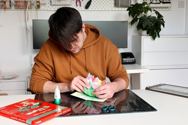 A young man sticks pop-up flowers to a card. There are glue, scissors and a pack of Canon paper on the surface next to him. 