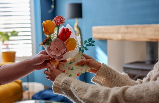 A homemade papercraft bouquet of flowers is passed from one pair of hands to another within a living room setting.  