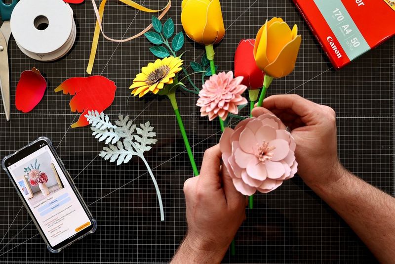 A pair of hands holds assembled papercraft flowers, with more placed on the table below. Next to them, a smartphone displays a picture of a papercraft bouquet on the Canon Creative Park app.
