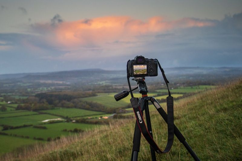 A Canon EOS R6 camera sits on a tripod on a grassy hillside, the fields below shown on the viewfinder.