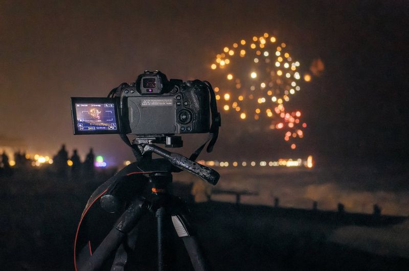 A Canon EOS R6 camera is set up on a tripod facing fireworks exploding over the sea.