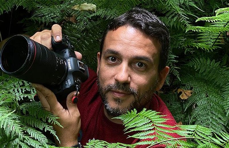 Canon Ambassador Joel Santos crouched among the ferns in a forest with his Canon camera. 