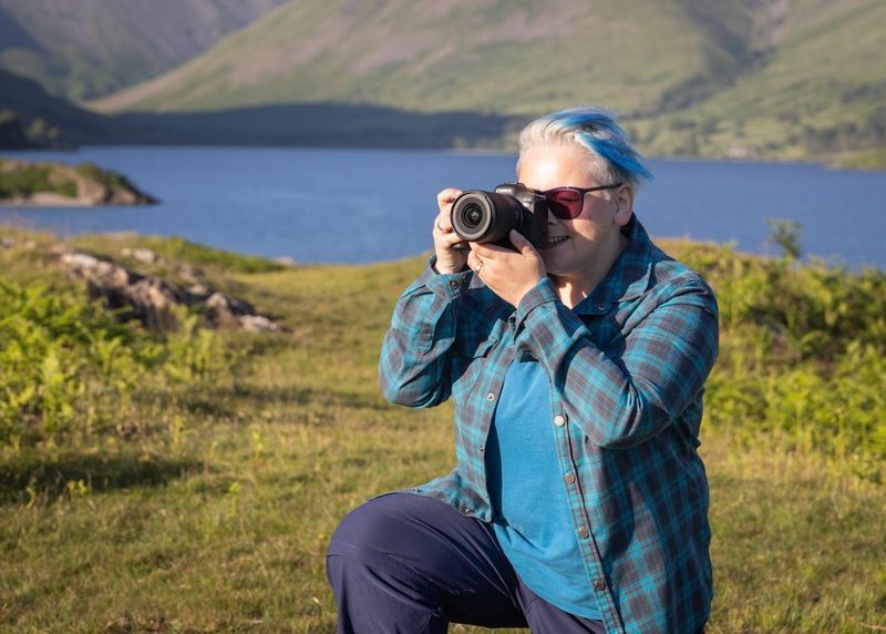 A female photographer holding a Canon camera kneels to take a photo. Behind her is a lake surrounded by mountains.