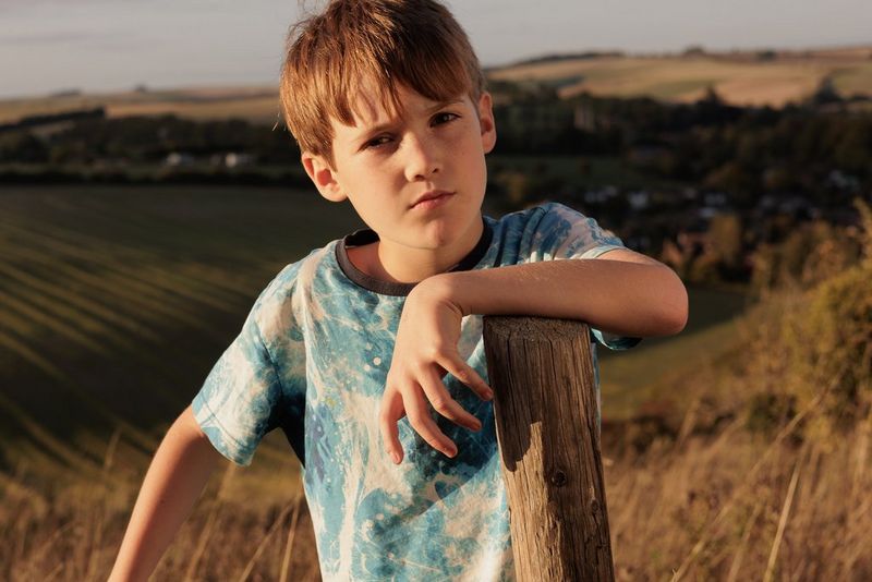 A child in a field resting his arm on a wooden post. A reflector has been used to soften the light. Taken on a Canon EOS R6 with a Canon RF 50mm F1.8 STM lens.